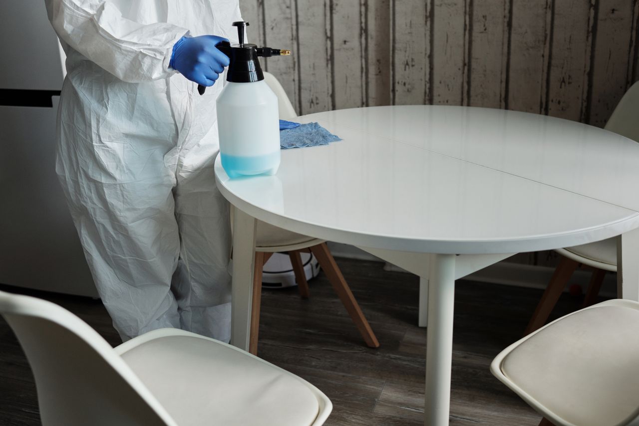 team-04 Person in protective suit disinfecting a white table indoors with a sprayer.