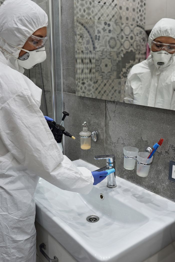who-we-are Person in protective gear disinfecting bathroom sink with spray bottle.